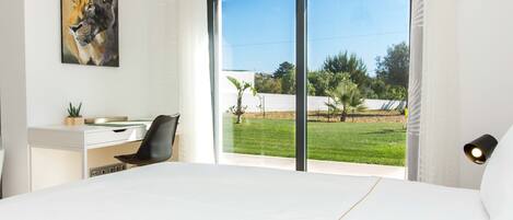 A bedroom with a large bed, a desk, a chair, a painting, and a view of a green landscape through a sliding door.