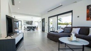 Un salon moderne avec un canapé, une table basse et un téléviseur. On aperçoit à l’arrière-plan un coin repas avec des chaises et une table.