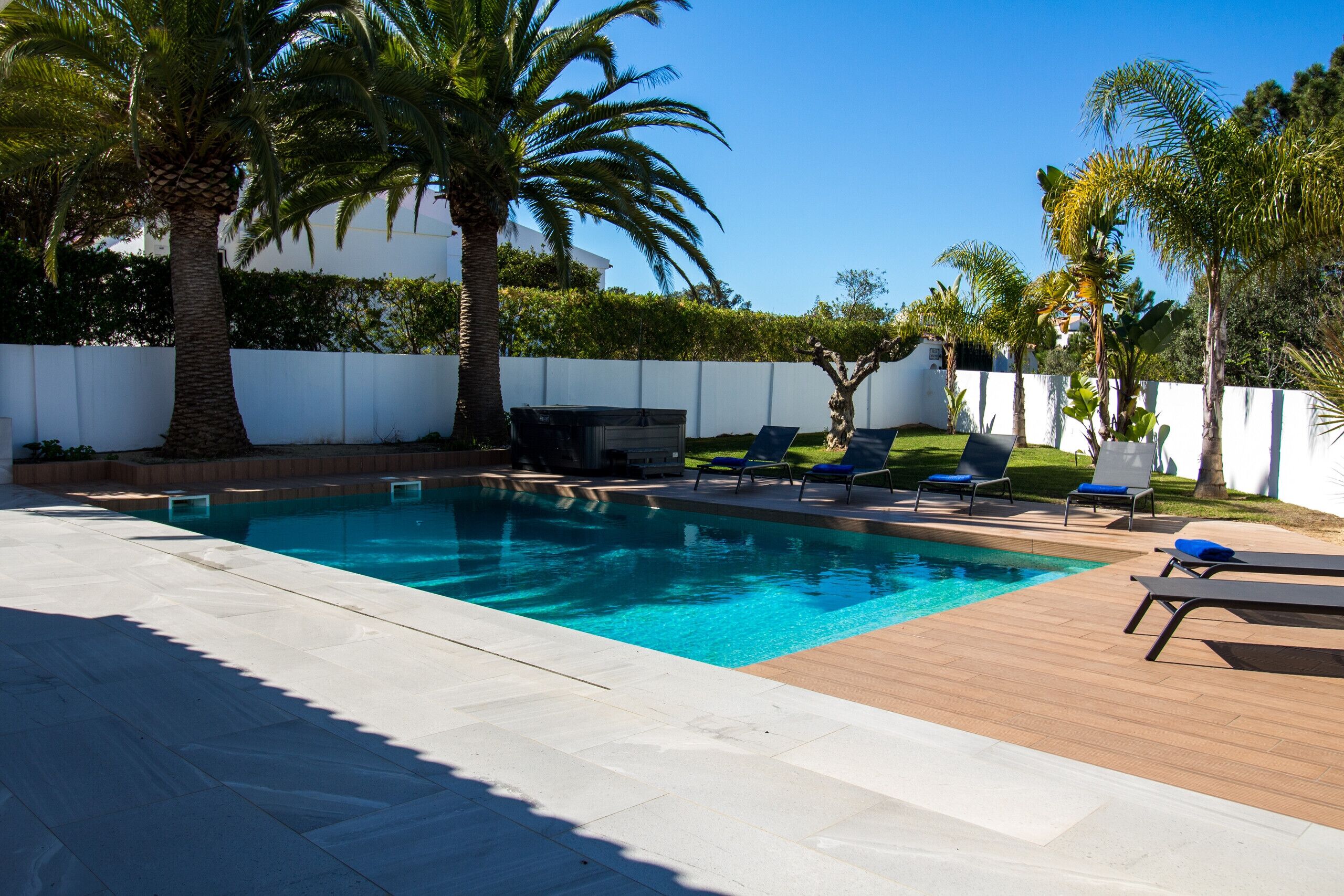 A rectangular swimming pool with clear blue water, surrounded by a wooden deck and white tiled flooring. There are several lounge chairs with blue cushions and a small outdoor grill area.