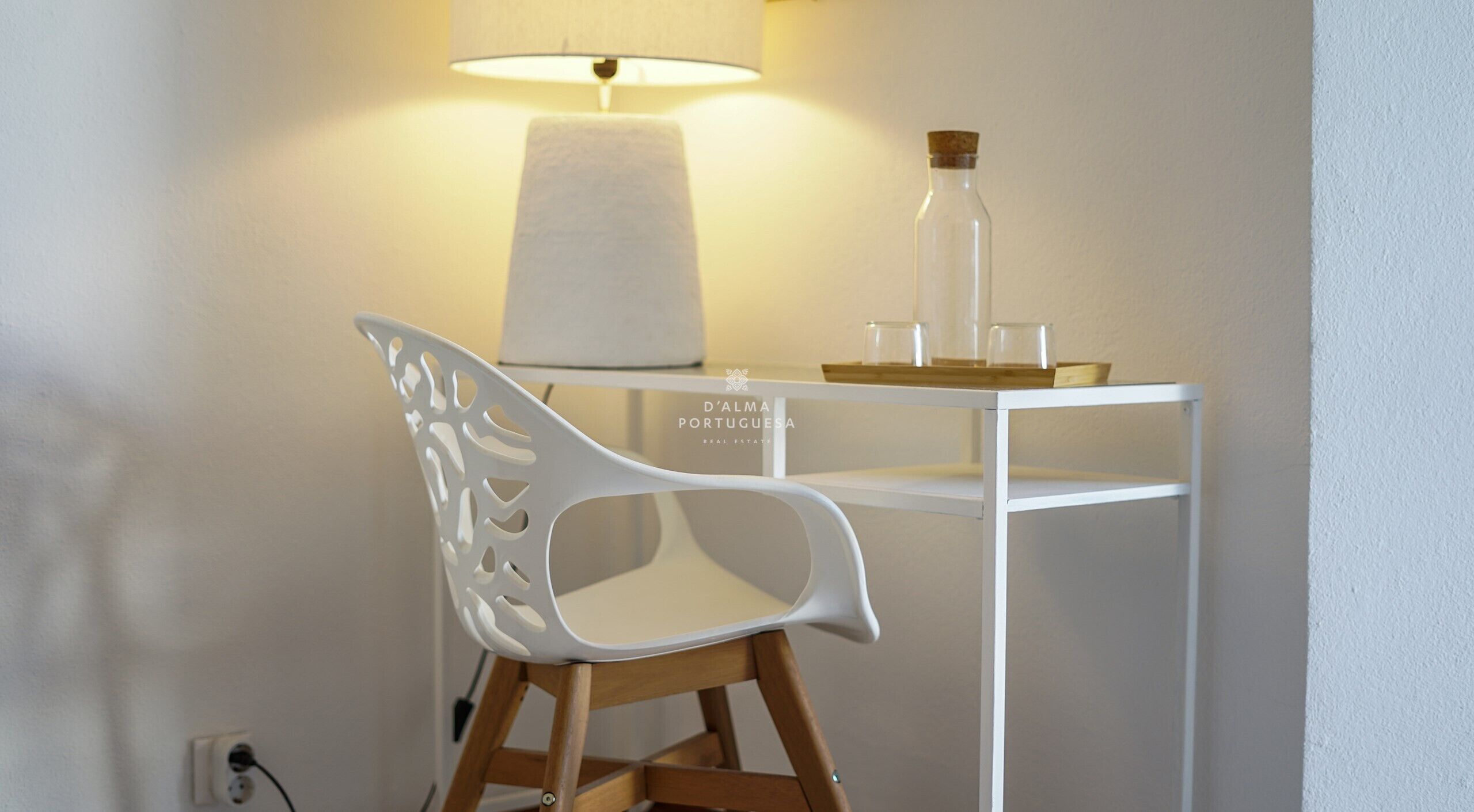 A white chair with a unique cut-out design, a white desk with a lamp, and a corked glass bottle on the desk.