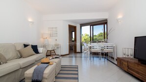 A modern living room with a sofa, coffee table, TV, dining area, and large windows. - Salgados Lounge Dalma Portuguesa (Albufeira)