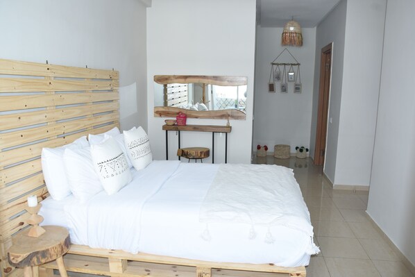 Une chambre à coucher avec une tête de lit en bois, un lit recouvert d’une literie blanche, une petite table ronde, un miroir et un luminaire suspendu.
