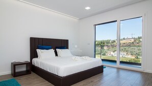 A modern bedroom with a large bed, a nightstand, a teal rug, and a balcony with a view of the landscape. - Dalma- Villa da Ourada (Albufeira)