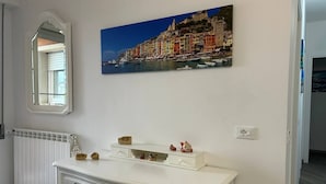 Een kamer met een witte kapstok, een spiegel, een radiator en een schilderij van een kustplaatsje.