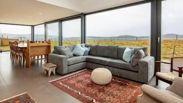 Un salon moderne avec un canapé d’angle, une table à manger et des chaises. De grandes fenêtres offrent une vue sur la campagne.