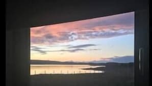 Une fenêtre donnant sur un coucher de soleil au-dessus d’une étendue d’eau et sur un paysage lointain.