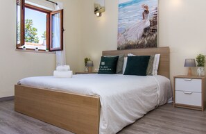 Un lit bien fait, avec un cadre en bois, une table de chevet avec une lampe, et un tableau représentant une femme au bord de la mer accroché au mur.