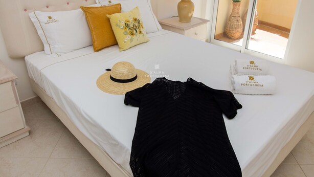 Un letto rifatto con una coperta bianca, un capo d'abbigliamento nero, un cappello di paglia e due asciugamani con un logo.