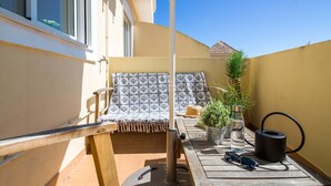 Un balcone con una tovaglia a motivi, una sedia, una pianta in vaso, una bottiglia d'acqua e un bollitore.