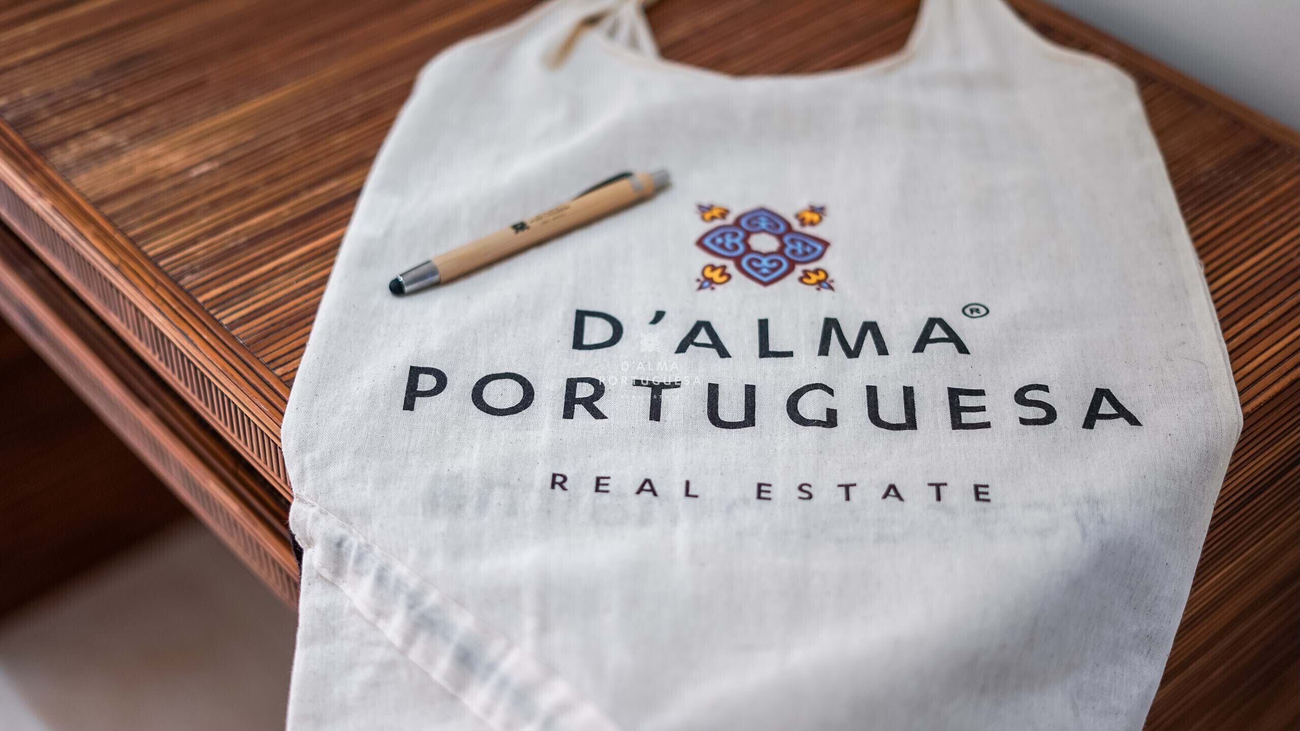 Un tablier blanc de la marque D'Alma Portuguesa, spécialisée dans l’immobilier, orné d’un logo et d’un texte, posé sur une surface en bois.