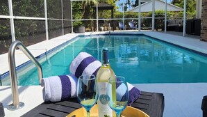 A poolside setting with a covered area, a pool, and a table with a bottle, glasses, and a bowl of fruit. - Naples Gem Near Beach | Heated Pool & BBQ (Naples)