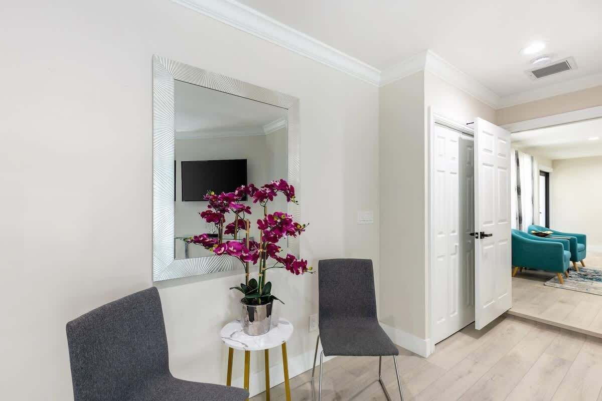 A hallway with a mirror, two chairs, a potted plant, and a door leading to a room with teal chairs.