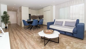 A modern living room with a blue sofa, a white coffee table, and a dining area with blue chairs. - Apart Terraços do Mar D´alma Portuguesa (Loulé)