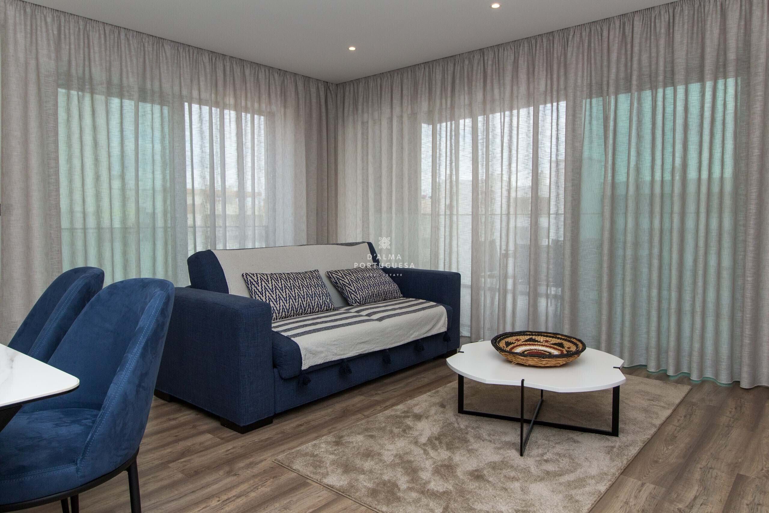 Ein modernes Wohnzimmer mit einem blauen Sofa, zwei Sesseln, einem weißen Couchtisch und einem großen Fenster mit durchsichtigen Vorhängen.