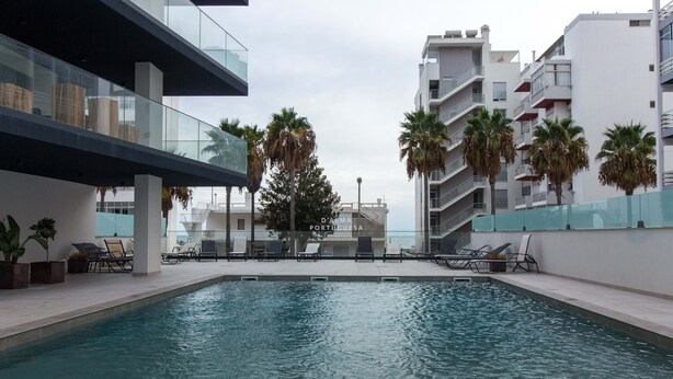 An indoor swimming pool with lounge chairs, palm trees, and modern buildings in the background. - Apart Terraços do Mar D´alma Portuguesa (Loulé)