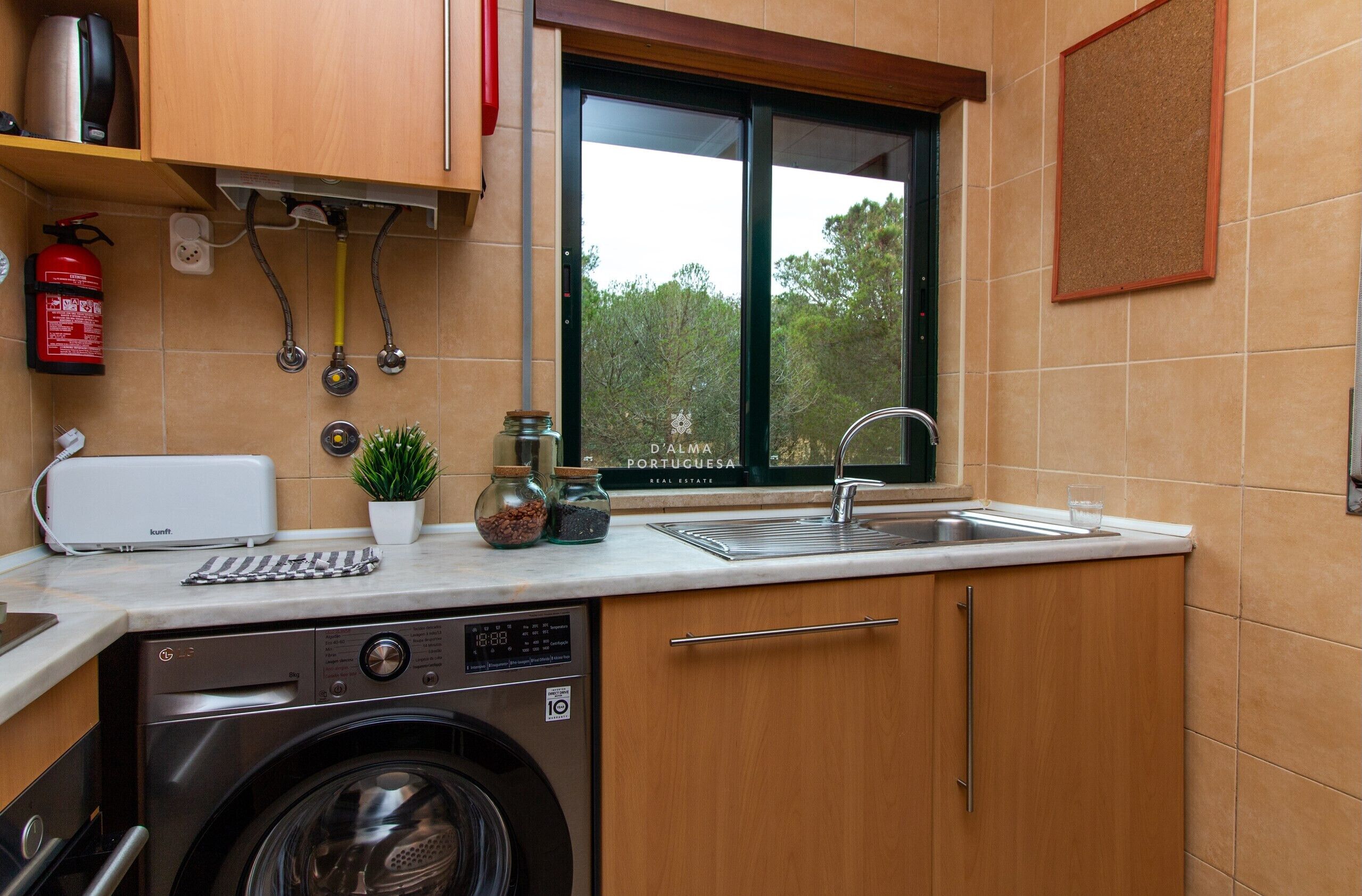 A compact kitchen with a washing machine, a toaster, a sink, and a window with a view of trees.