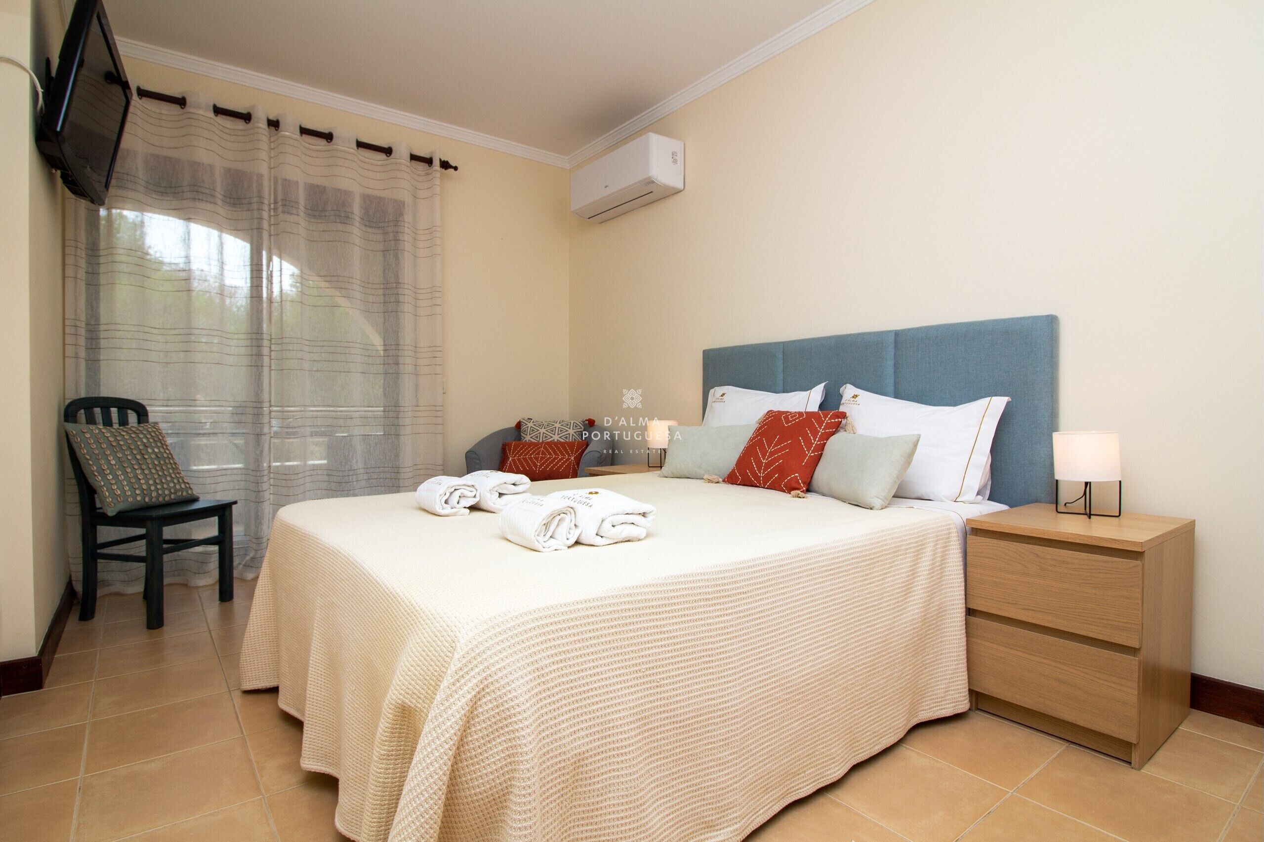 A neatly made bed with a blue headboard, a bedside table with a lamp, and a chair with a cushion.