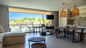 Una sala moderna con un sofá, una mesa de centro y un comedor con sillas y una mesa. Un televisor está montado en la pared y hay un balcón con vistas a palmeras.