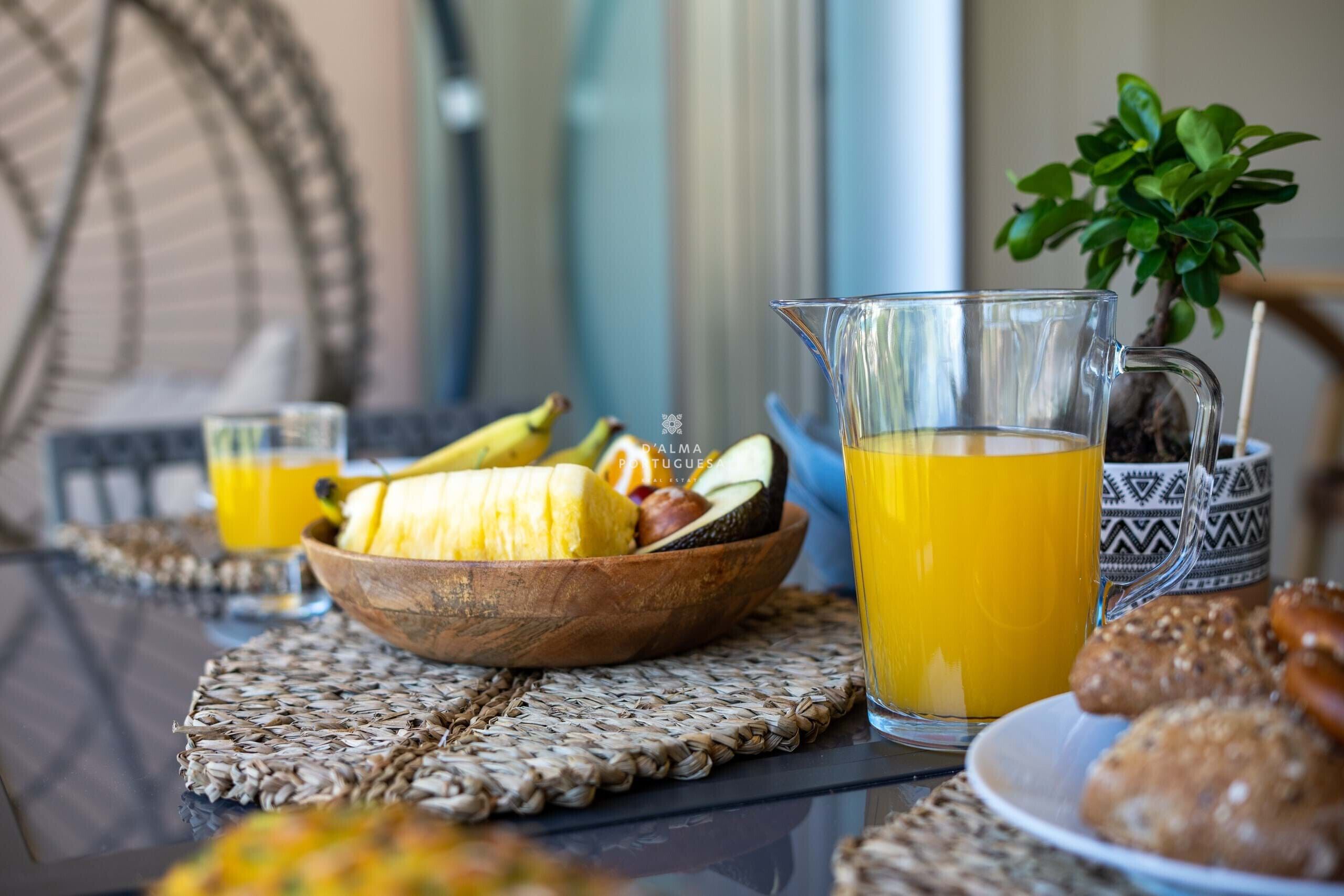 Ein Frühstückstisch mit einer Hollandschale voll Obst, einer Karaffe Orangensaft und einem kleinen Glas Orangensaft auf einer gewobenen Tischmatte.