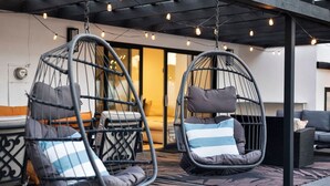 Deux fauteuils suspendus avec coussins, un tapis à motifs et un plafond moderne avec des poutres apparentes et des guirlandes lumineuses.