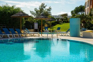 Piscina com espreguiçadeiras e guarda-sóis, cercada por vegetação e um edifício ao fundo.