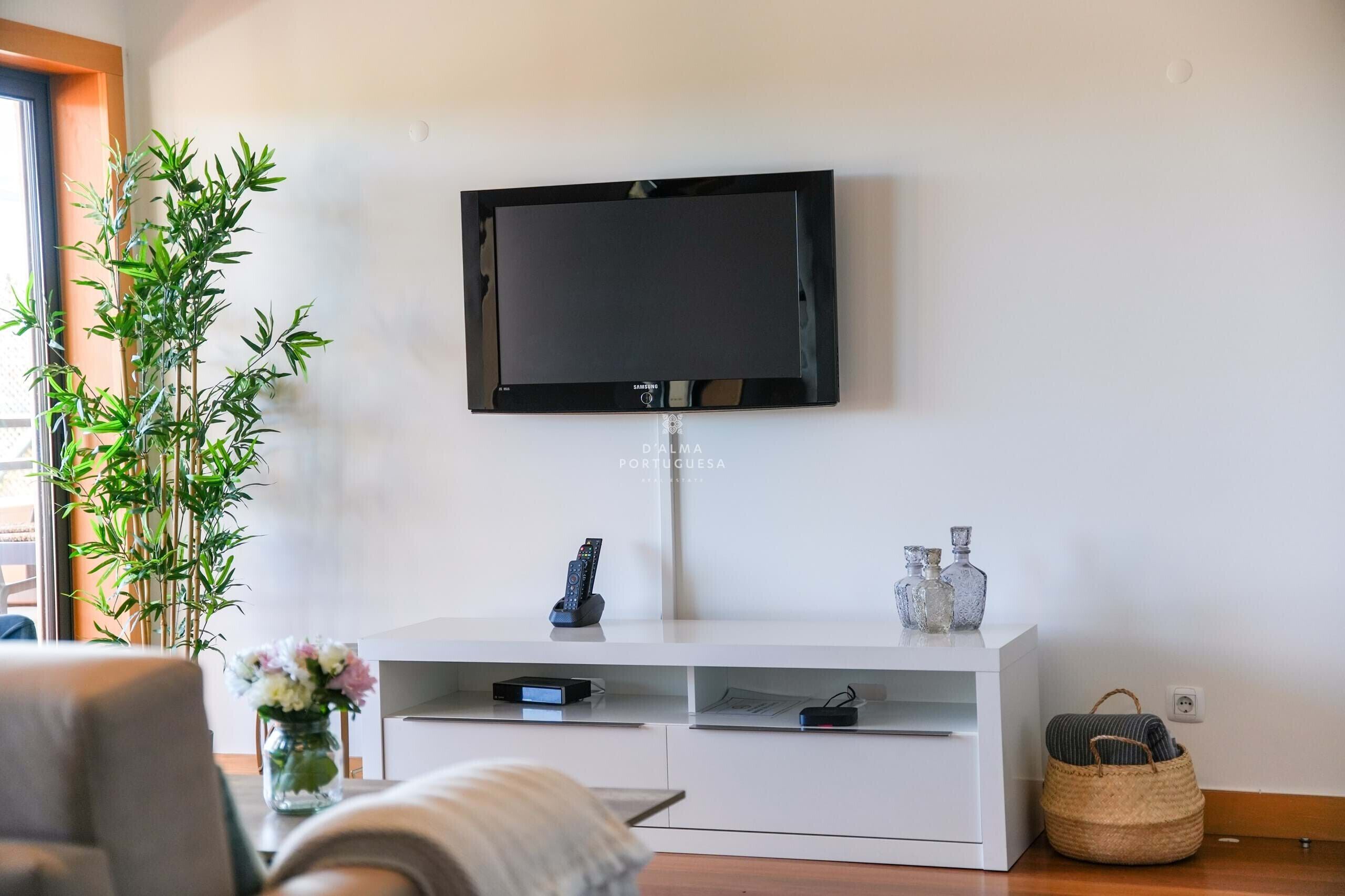 Un salon avec un téléviseur fixé au mur, un meuble de rangement blanc, une plante en pot et un panier en osier.