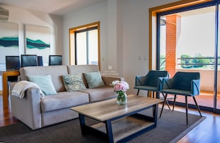 Uma sala de estar moderna com um sofá, uma mesa de centro e duas cadeiras. Há duas janelas com vista para o exterior.