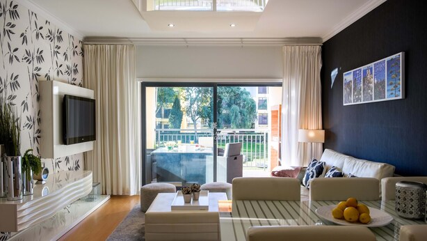 A modern living room with a flat-screen TV, comfortable seating, a glass coffee table, and a balcony with outdoor furniture. - Apart Vilamoura Pine D'alma Portuguesa (Loulé)