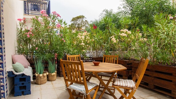 Een terras buiten met houten meubels, waaronder een tafel en stoelen, omgeven door potplanten en bloemen.
