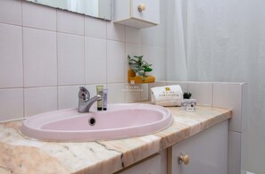 Egy fürdőszoba, amelyben rózsaszín mosdókagyló, tükör és egy polc található, amin a tisztálkodási cikkek vannak.