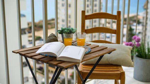 Ein Holztisch mit einem Stuhl, einem Buch, Brillen und einer kleinen Pflanze auf einem Balkon mit Blick auf Gebäude und das Meer.