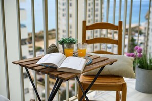A wooden chair and table with a book, glasses, and a small plant on a balcony with a view of buildings and the sea. - Apartamento Canela by D'alma Portuguesa (Silves)