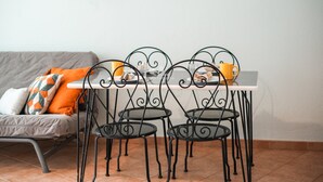 En matplats med ett vitt bord och fyra svarta metallstolar. En grå soffa med orange och vita kuddar står längs väggen.