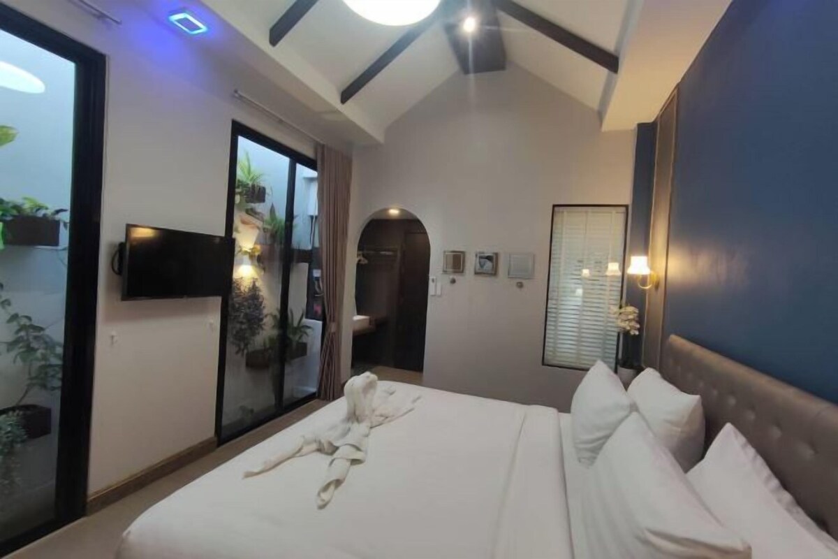 A hotel room with a bed, a TV mounted on the wall, and a sliding glass door leading to a balcony with plants.