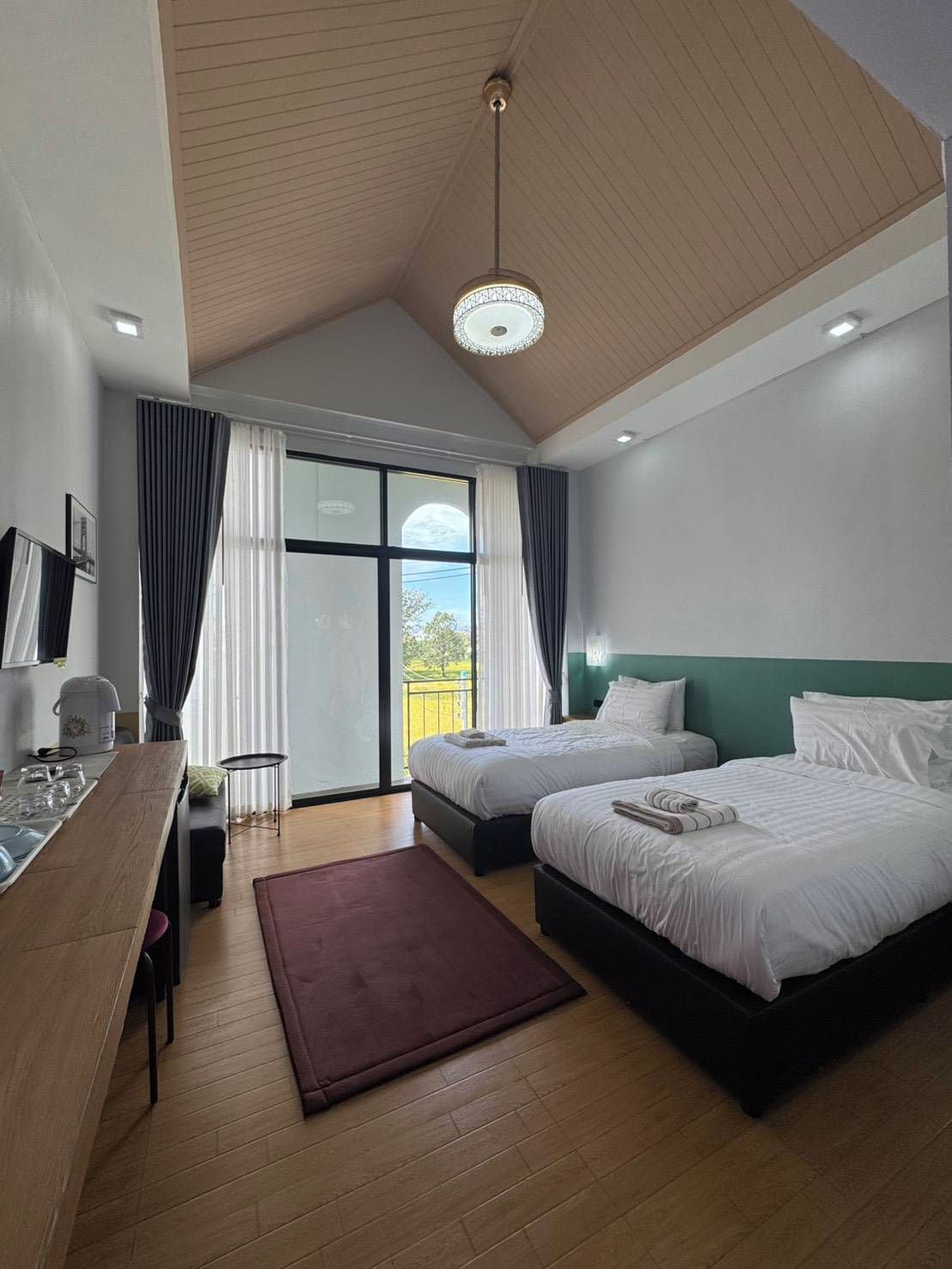 A hotel room with two beds, a dining area, a view through the window, and a ceiling light.