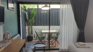A hotel room with a bed, a desk with a chair, a small table, and a potted plant. There is a sliding glass door leading to an outdoor area with a table and chairs. - U-SABAI VILLA  (Yasothon)