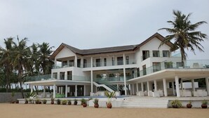 Una propiedad frente a la playa con varios pisos, un balcón grande, palmeras y una playa arenosa.