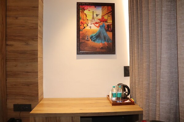 Une chambre d’hôtel avec un lambris en bois, un tableau encadré représentant une femme en robe bleue, et une table d’appoint en bois sur laquelle se trouvent une bouilloire et des bouteilles.