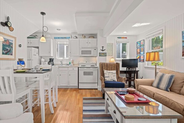 A modern kitchen with white cabinetry, a dining area with white chairs, a sofa, and a TV. - Private Deck, Parking, Walk to Town! (Provincetown)