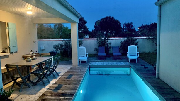 Uma área de piscina com deck de madeira, uma mesa com cadeiras e duas espreguiçadeiras.