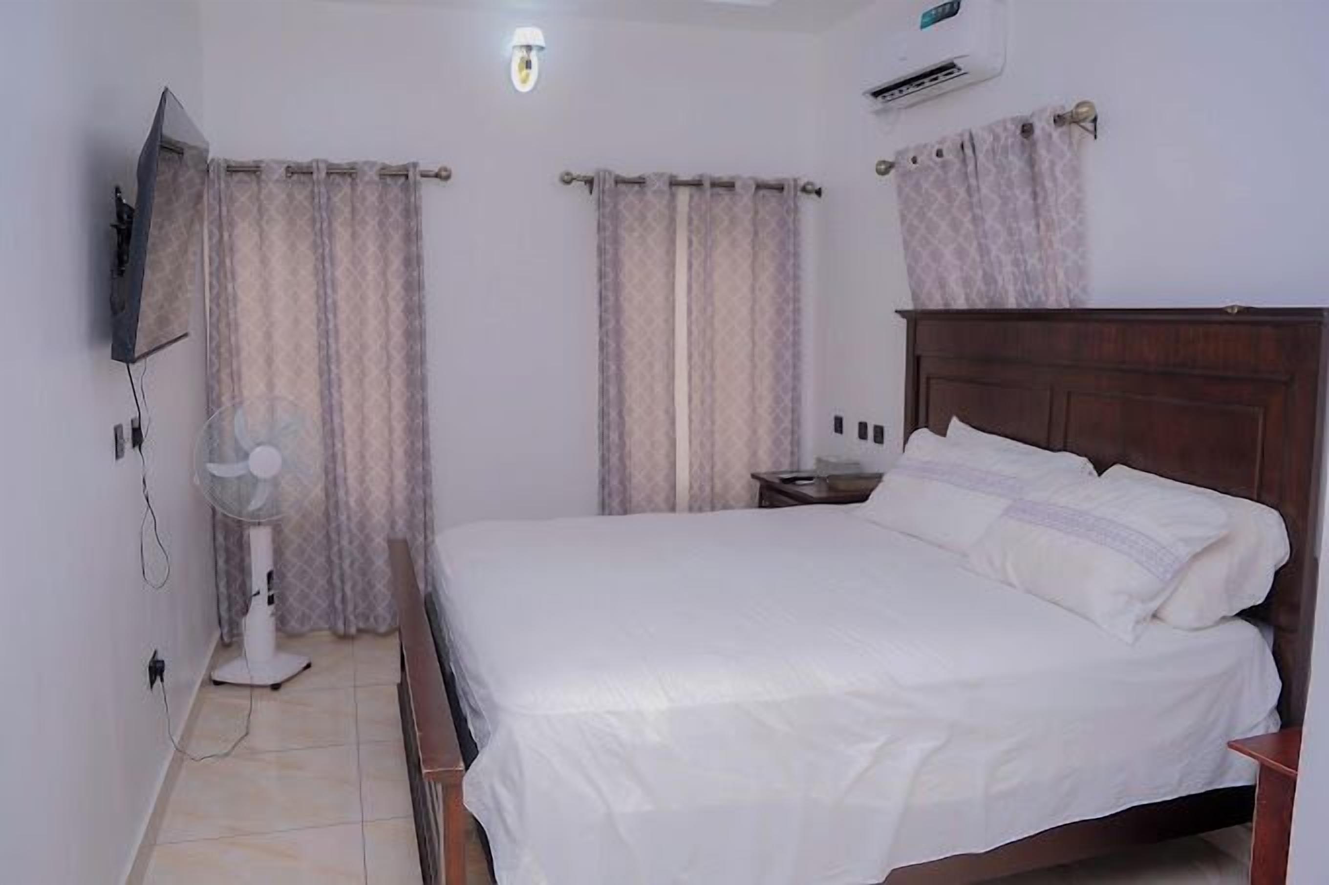 A bedroom with a bed, a bedside table, a wall-mounted TV, a ceiling fan, and curtains.