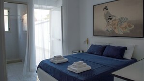 Ein Hotelzimmer mit Bett, Nachttisch, Wandklimaanlage, Fenster mit durchsichtigen Vorhängen und einem Bild an der Wand.