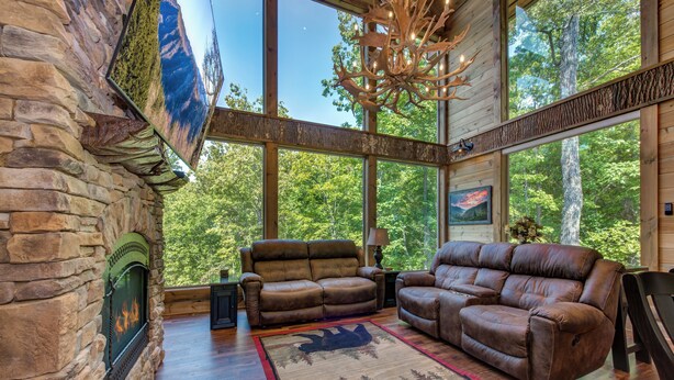 Ein Blockhaus-Interieur mit Steinwänden, einem Kamin, Ledersofas und großen Fenstern mit Blick auf einen Wald.