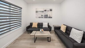 En moderne stue med to sorte sofaer, et træbord og et stort vindue med persienner.