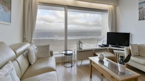 A living room with a large window overlooking the beach, a sofa, a coffee table, and a TV. - Flat in Blankenberge With Ocean Views (Blankenberge)