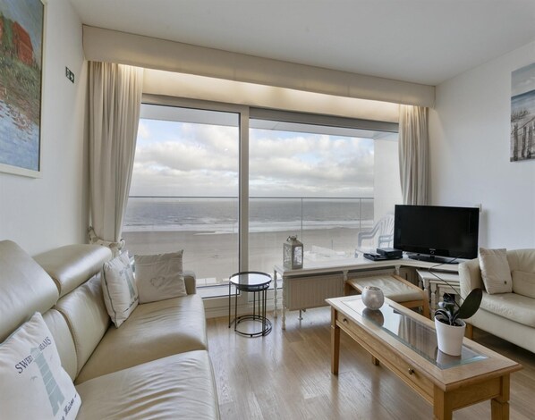 A living room with a large window overlooking the beach, a sofa, a coffee table, and a TV. - Flat in Blankenberge With Ocean Views (Blankenberge)