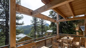 Eine Holzterrasse mit Tischen und Bänken, die auf einen bewaldeten Bereich mit Bergen im Hintergrund blickt.