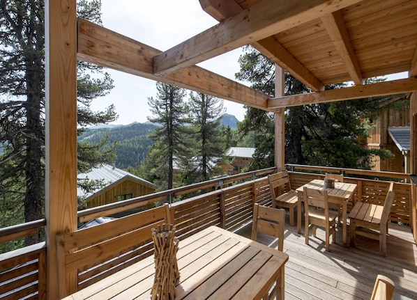 A wooden deck with tables and benches, overlooking a forested area with mountains in the background.