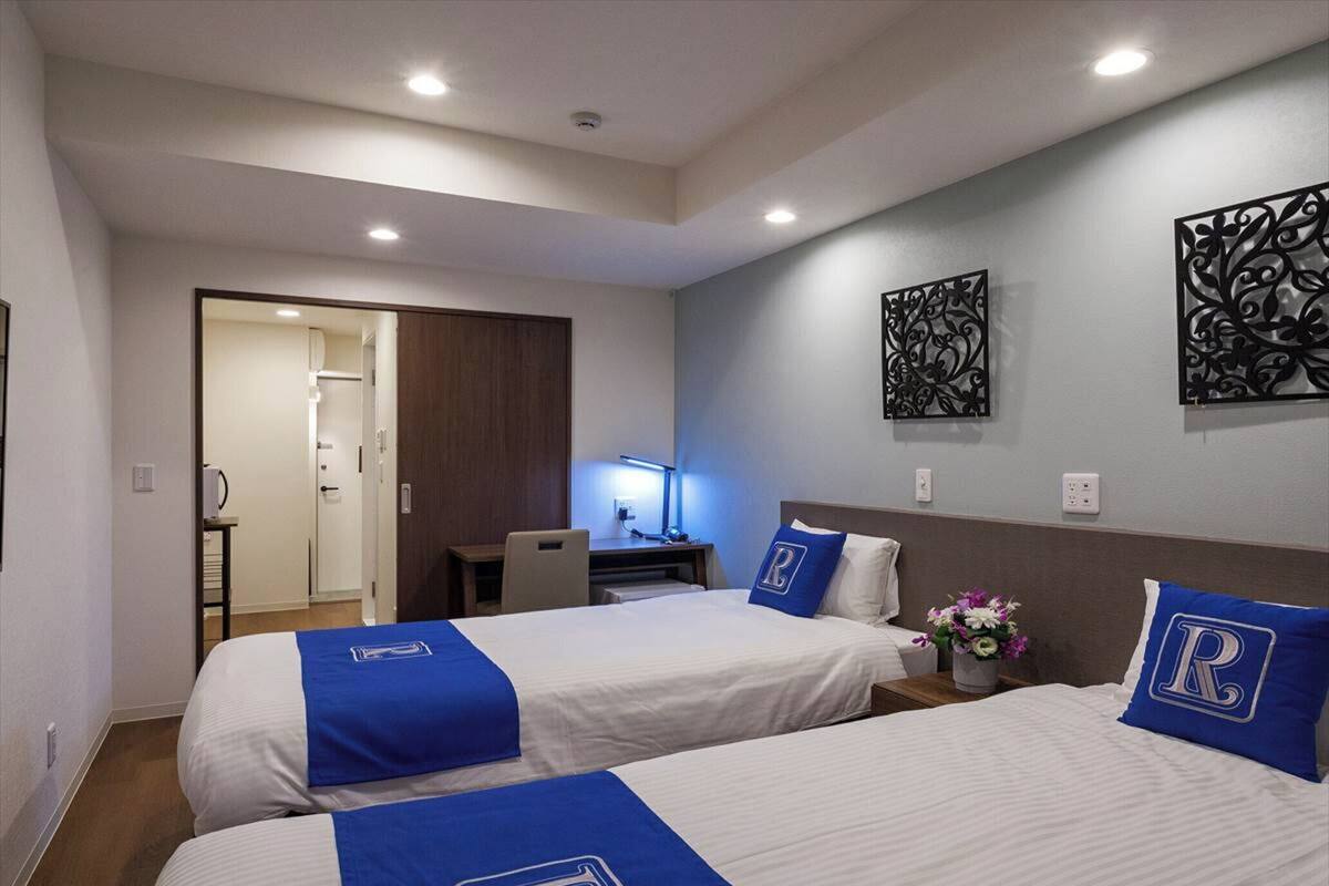 A hotel room with two beds, each with a blue bedspread and a pillow with a 'P' logo. There is a desk with a lamp, a wall-mounted decorative piece, and a door leading to another room.