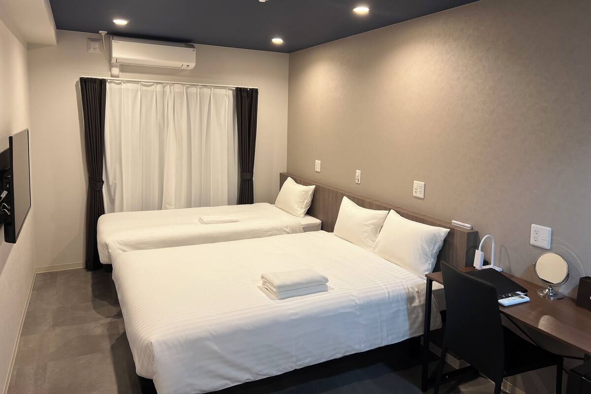 A hotel room with two beds, a desk, a chair, a TV, and an air conditioning unit.